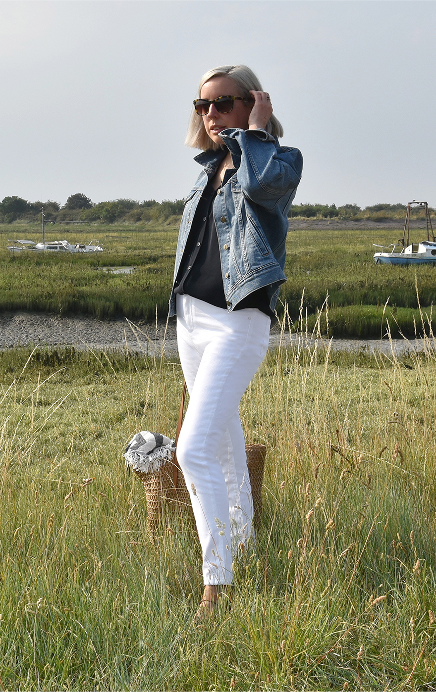 Jen Wilkins, Creative Copy & Editorial Manager at Hobbs stands in a secluded spot by the coast wearing a blue denim jacket, a navy blue blouse, white denim jeans and tan sandals styled with a straw bag.